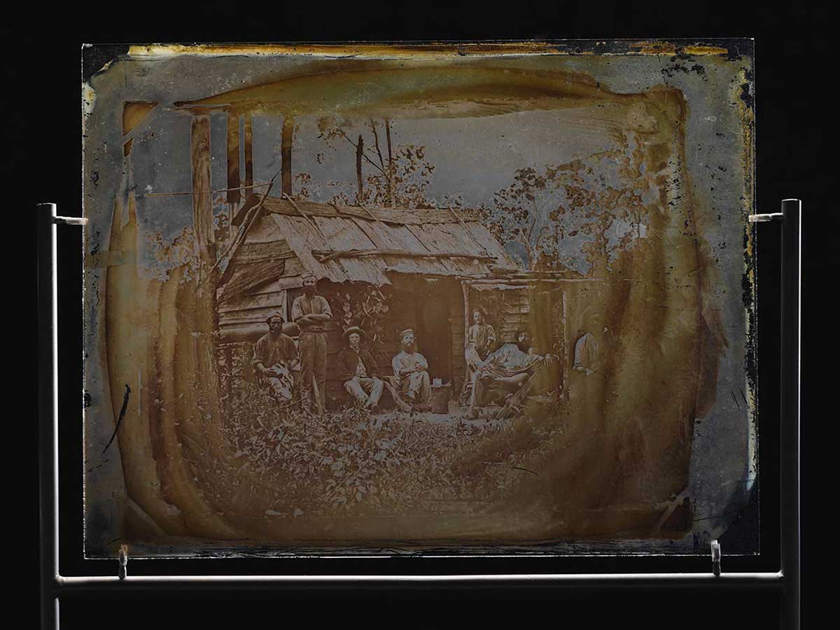 Glass showing seven men outside a bush slab hut. Most of the men are sitting and the man on the far right is only partially visible. - click to view larger image