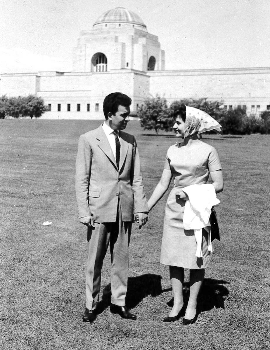 A black and white photo of a man and woman holding hands in. They are outdoors, standing on a lawn with a large white building in the background. - click to view larger image