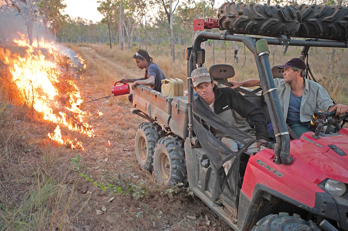 Three people in an all-terrain vehicle in the bush. A man at the back sets alight grass by the roadside.
