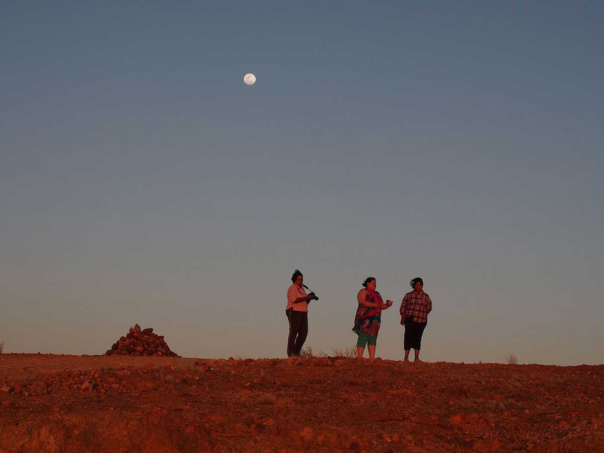 Long distance view of three people standing in an arid landscape of low scrub and rocky terrain. - click to view larger image