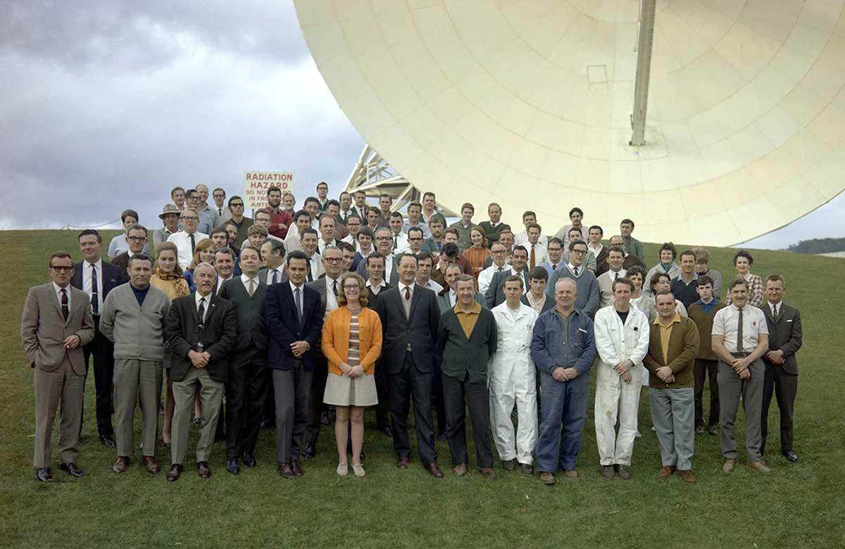 A group portrait of men and women standing in front of a satellite dish.