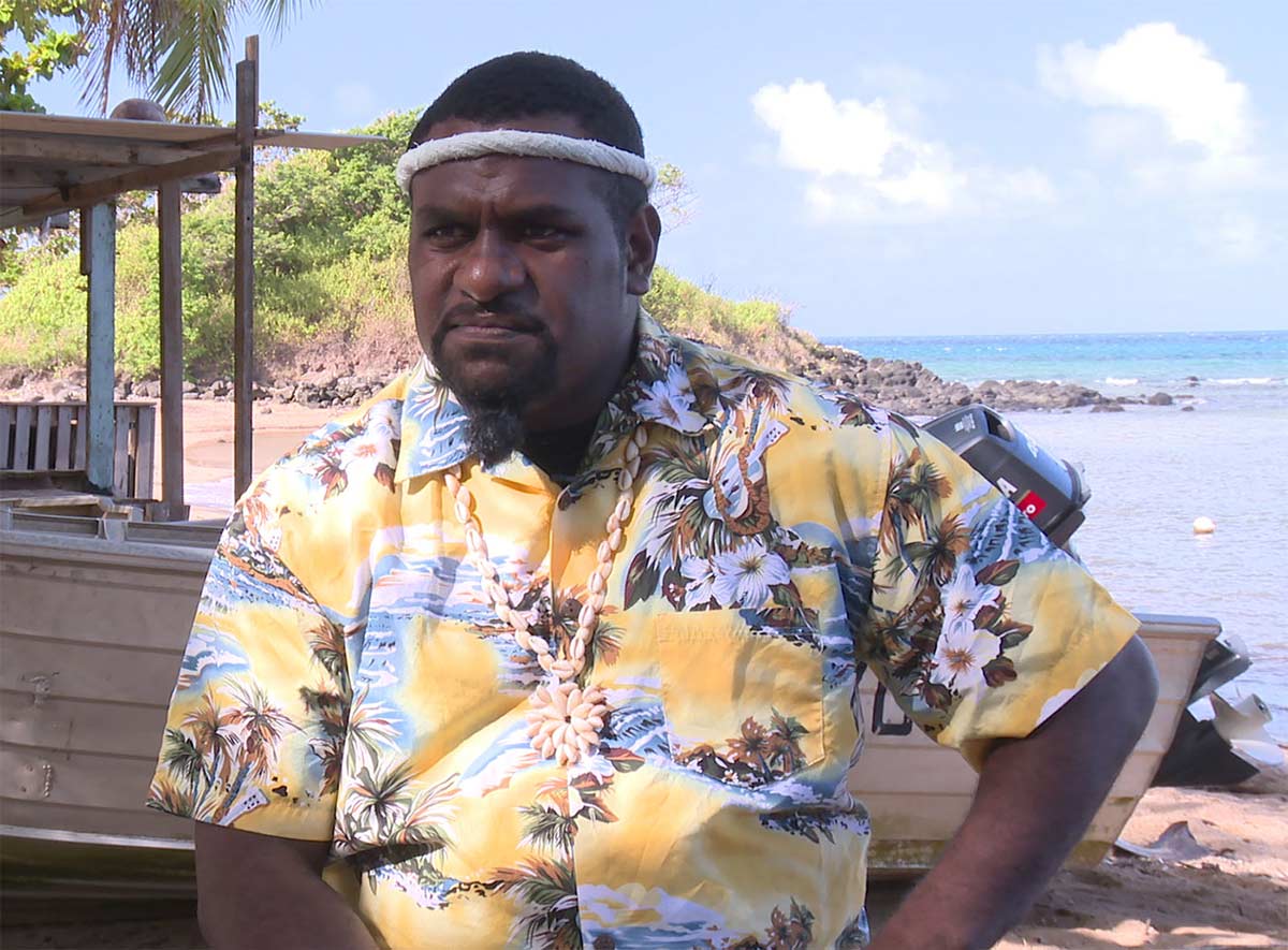 Photo of a man on a beach and wearing a white headband and a shirt with tropical motif.