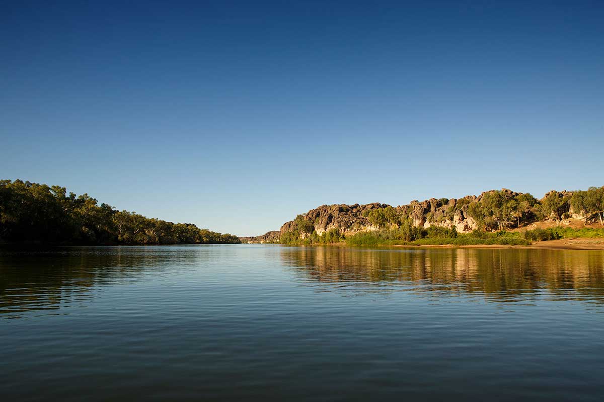 View of a river running through sheer ochre rock formations on one side and trees on the other.