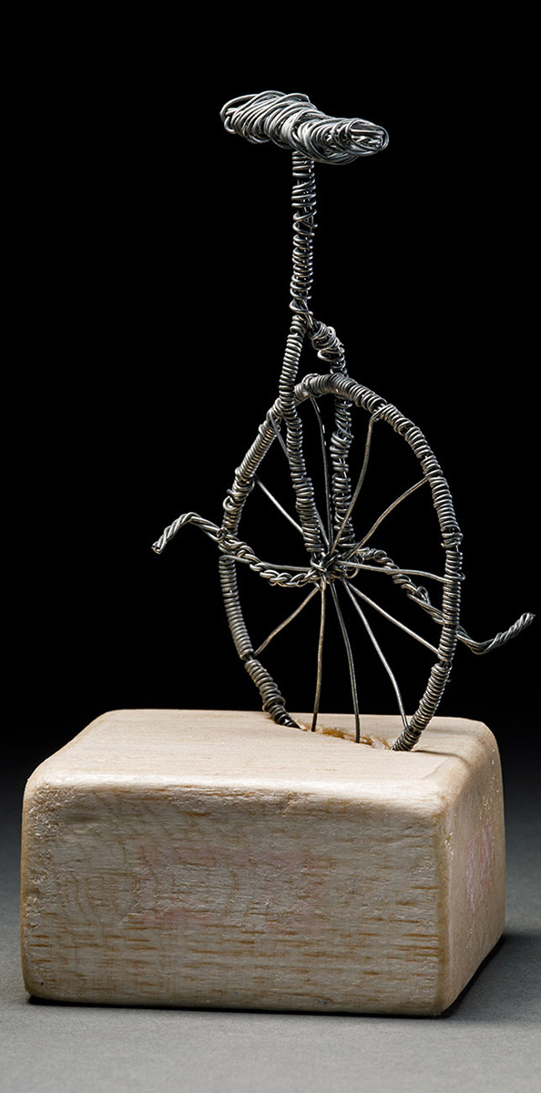 A handmade trophy that is composed of a miniature wire sculpture of a unicycle that sits in a diagonal slot carved from a block of light coloured balsa wood. - click to view larger image