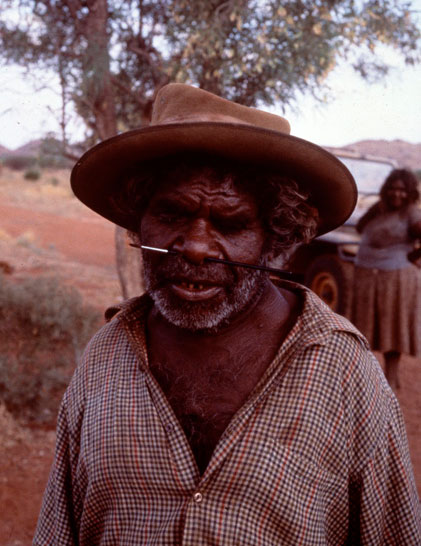 Portrait photo of an Aboriginal Australian man in a hat with a paintbrush through his nose. - click to view larger image