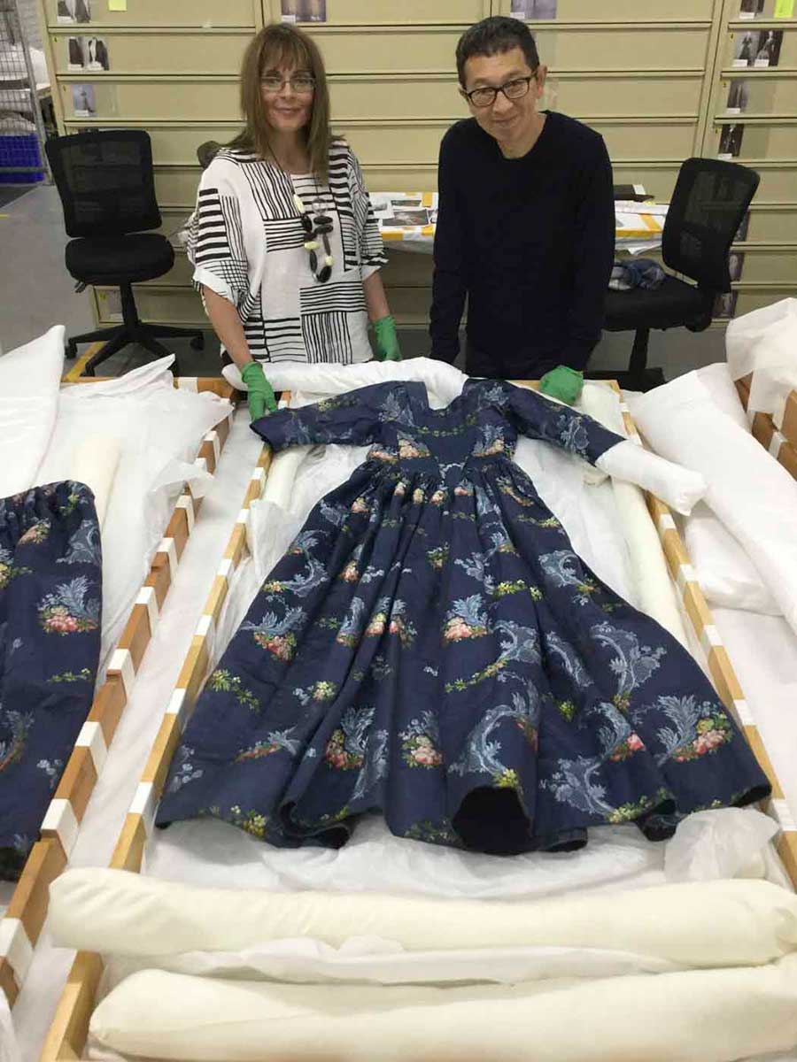 Two conservators stand behind a dark blue dress which is displayed on a bench. - click to view larger image