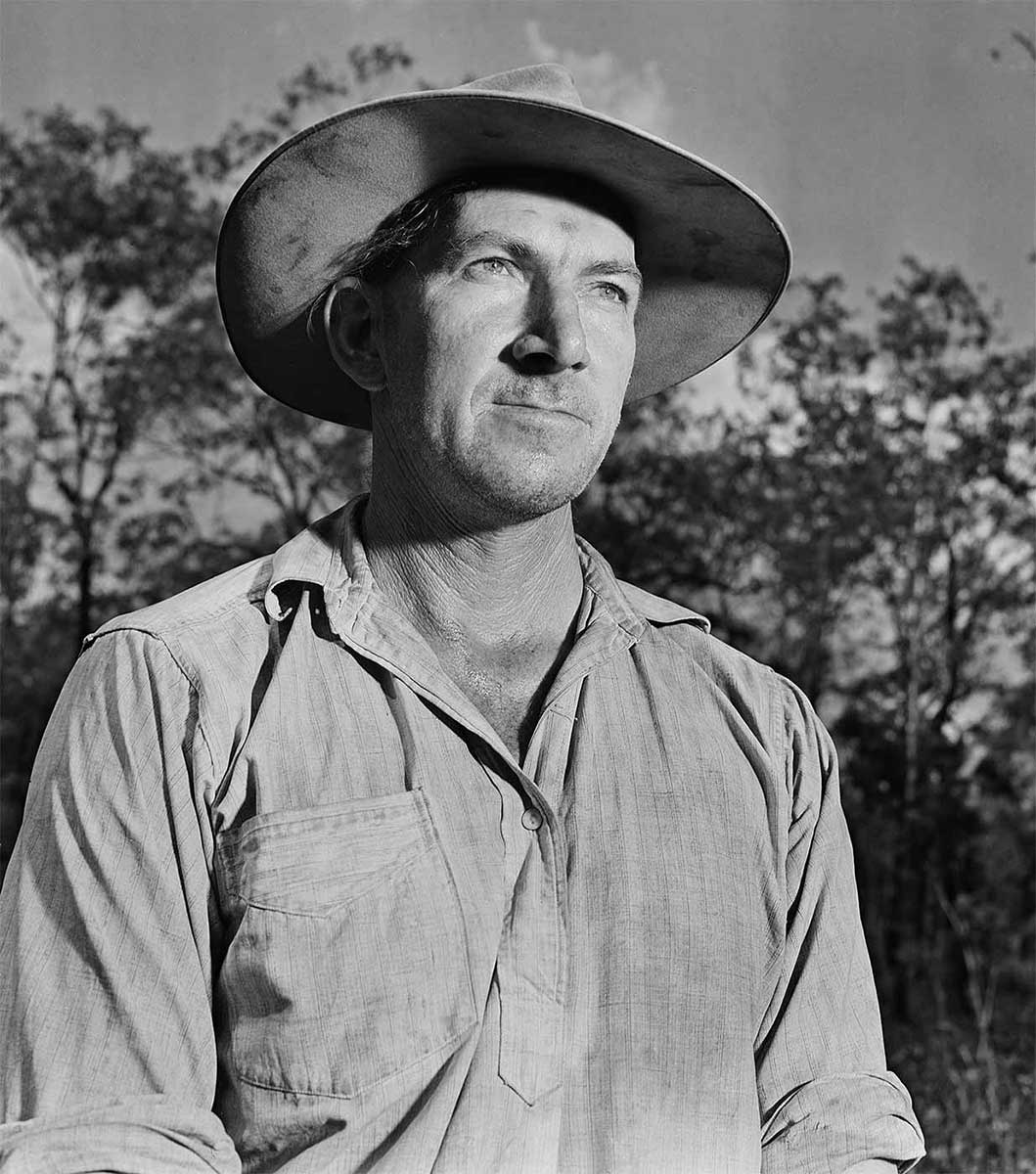 Black and white portrait of a man wearing an Akubra hat and an old shirt. He is looking off into the distance and there is a blurred image of a forest in the background. - click to view larger image