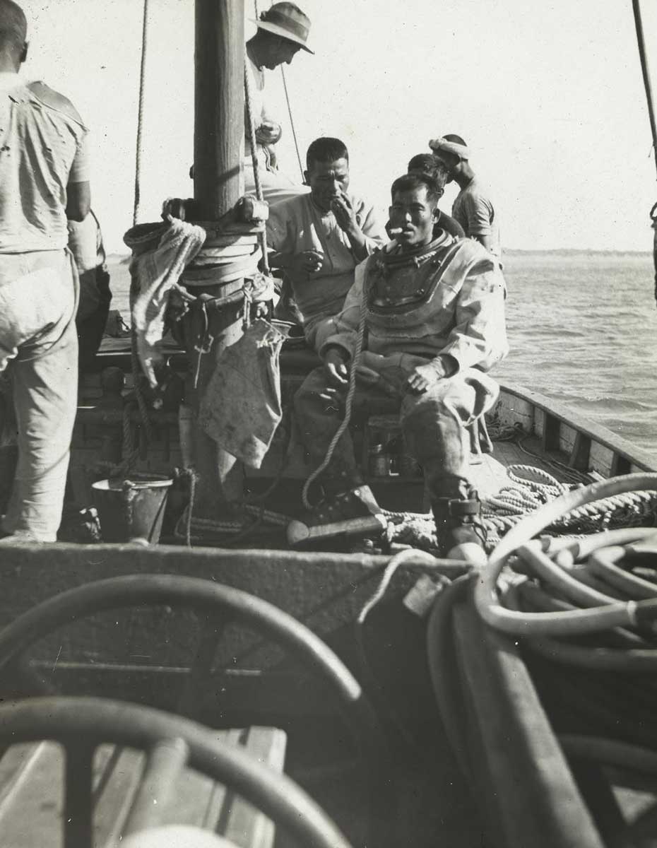 Black-and-white photo of a small group of men sitting and standing on the deck of a sailing vessel. - click to view larger image