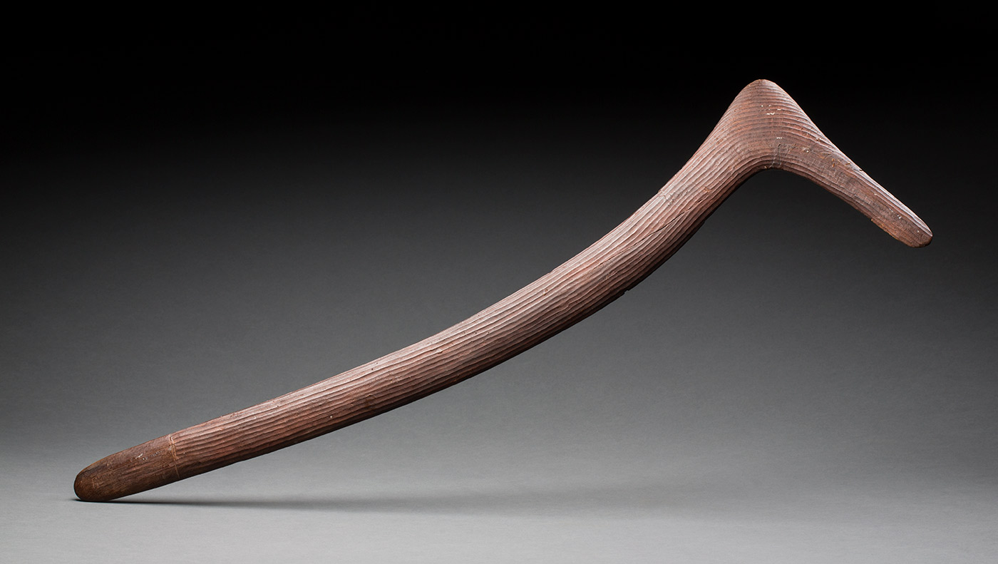 A brown wooden boomerang that is asymmetrical. It is tick shaped with one long side and one short side. It has a crack at the shorter end that has been repaired. One side of the boomerang is smooth and the other is decorated with carved lines. - click to view larger image