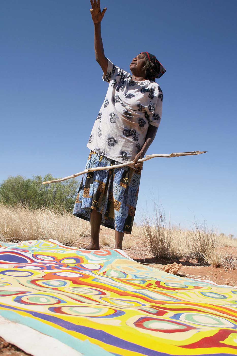 Colour photograph of a woman holding a stick with her arm raised towards the sky. A painting is lying on the ground at her feet. - click to view larger image