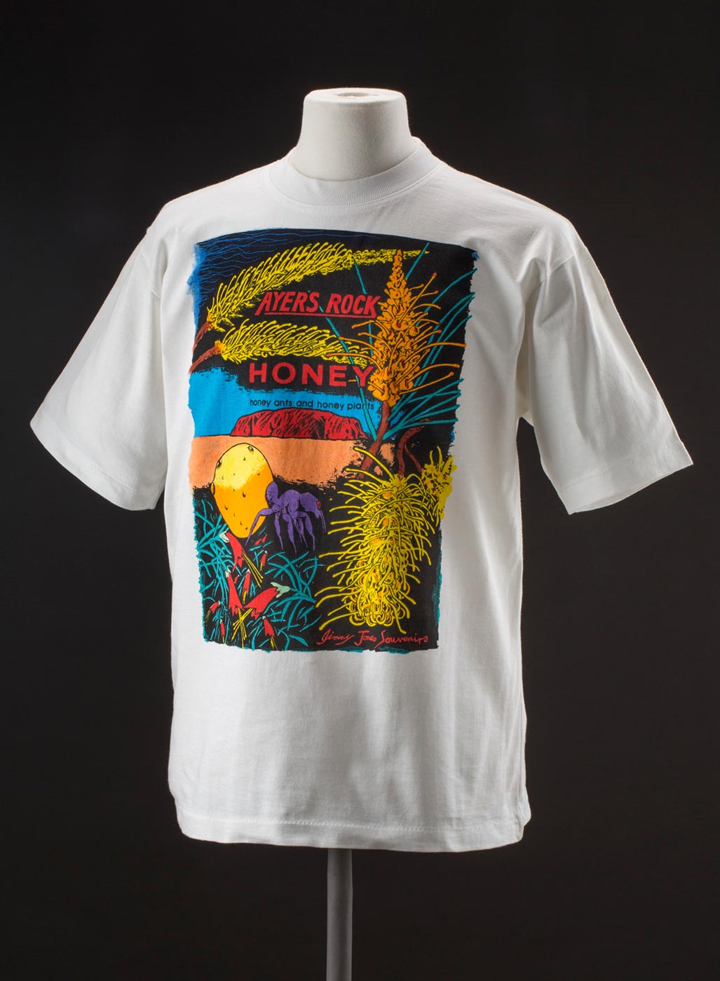 Depicted on the T-shirt is a brightly coloured landscape featuring native flowers and honey ant in foreground, Uluru / Ayers Rock in background and text above 