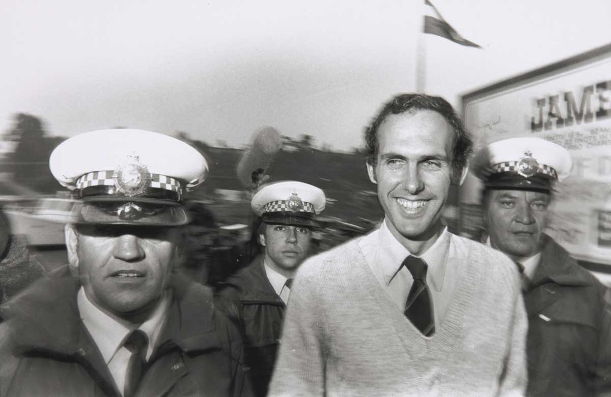A black and white photograph of Bob Brown surrounded by three police men. Bob is smiling and the officers look stern. A boom microphone can be seen in the back ground as well as a flag on a flag pole next to a bill board.