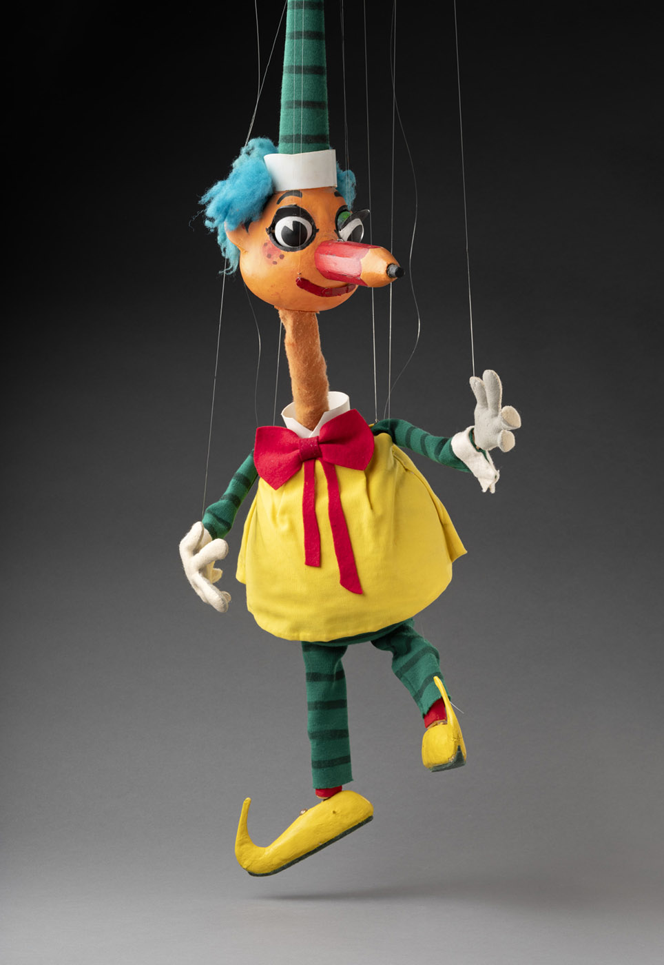 Mr Squiggle puppet with blue hair and a pencil for a nose. He is wearing a yellow pinafore with a red bow around the neck, green and black striped sleeves and leggings. He has white gloves, and yellow clog-like shoes that curve at the toes. His nose is a large red pencil with a piece of black oil pastel inserted at one end. Attached to his head is a long tapered green and black striped hat.