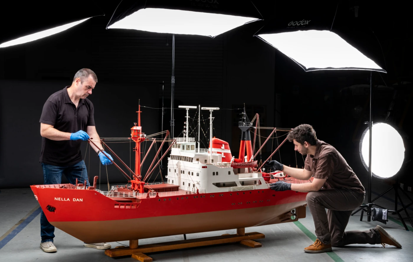 Two people in a photography studio beside a large model ship bright red on top. There are several large studio lights in place, illuminating the model.,