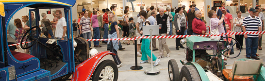 Visitors in a museum storage area looking at a vintage vehicle and a small tractor.